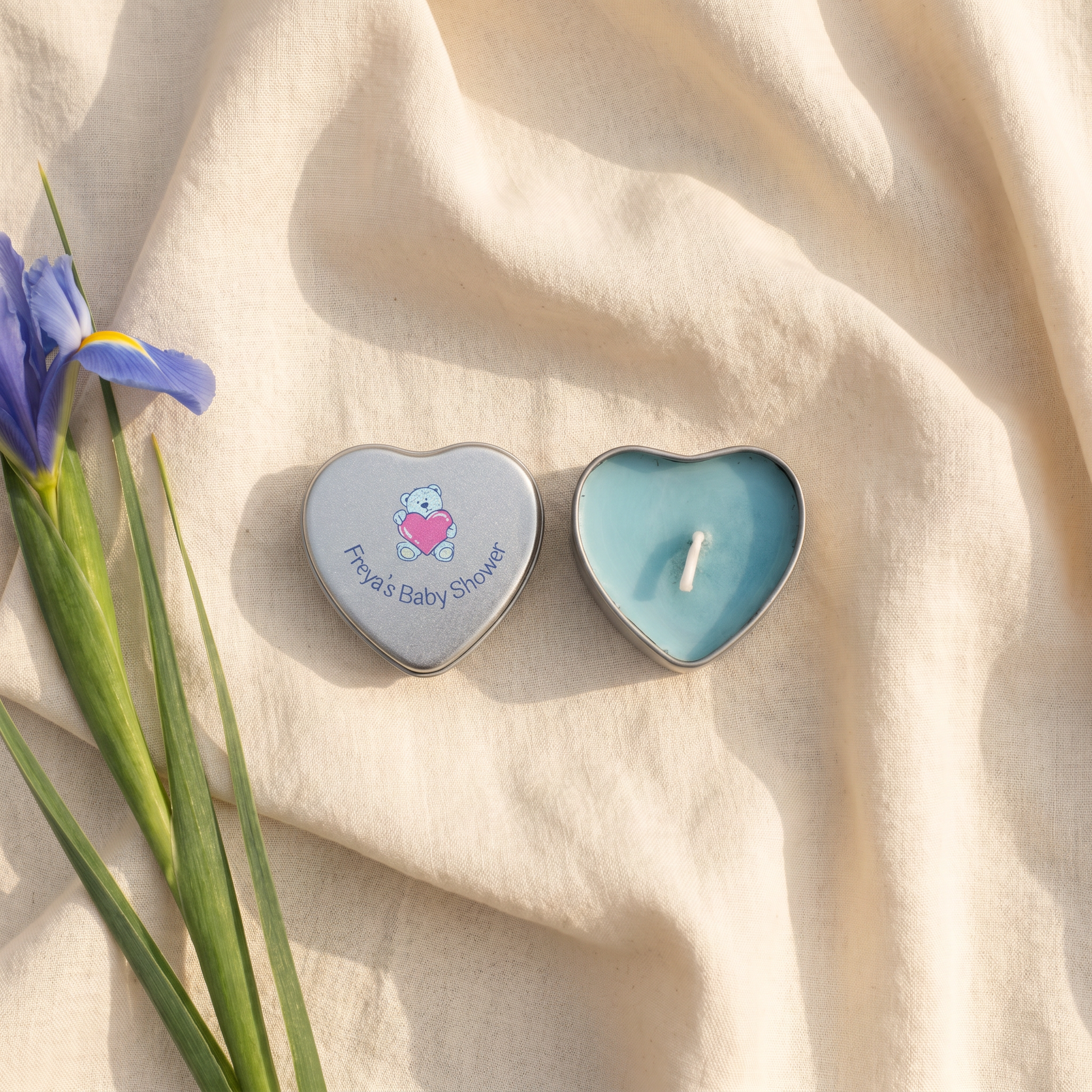 Personalised blue baby shower candle favour — silver heart tin open showing blue wax candle with teddy bear lid design, styled with iris flowers