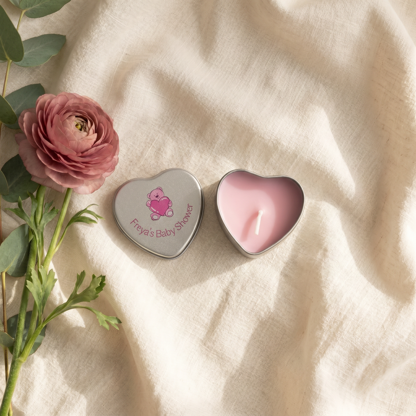 Personalised pink baby shower candle favour — silver heart tin open showing pink wax candle with teddy bear lid design, styled with pink ranunculus flowers