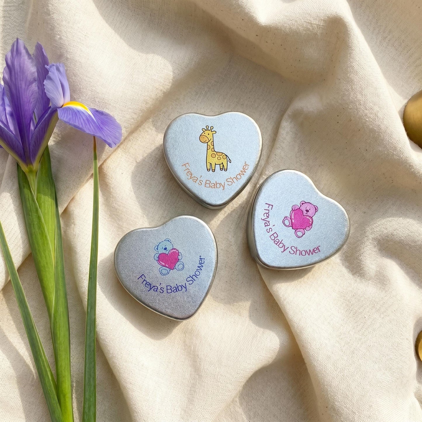 Three personalised baby shower heart tin candle favours showing lid designs — blue teddy bear, giraffe and pink teddy bear, with iris flowers