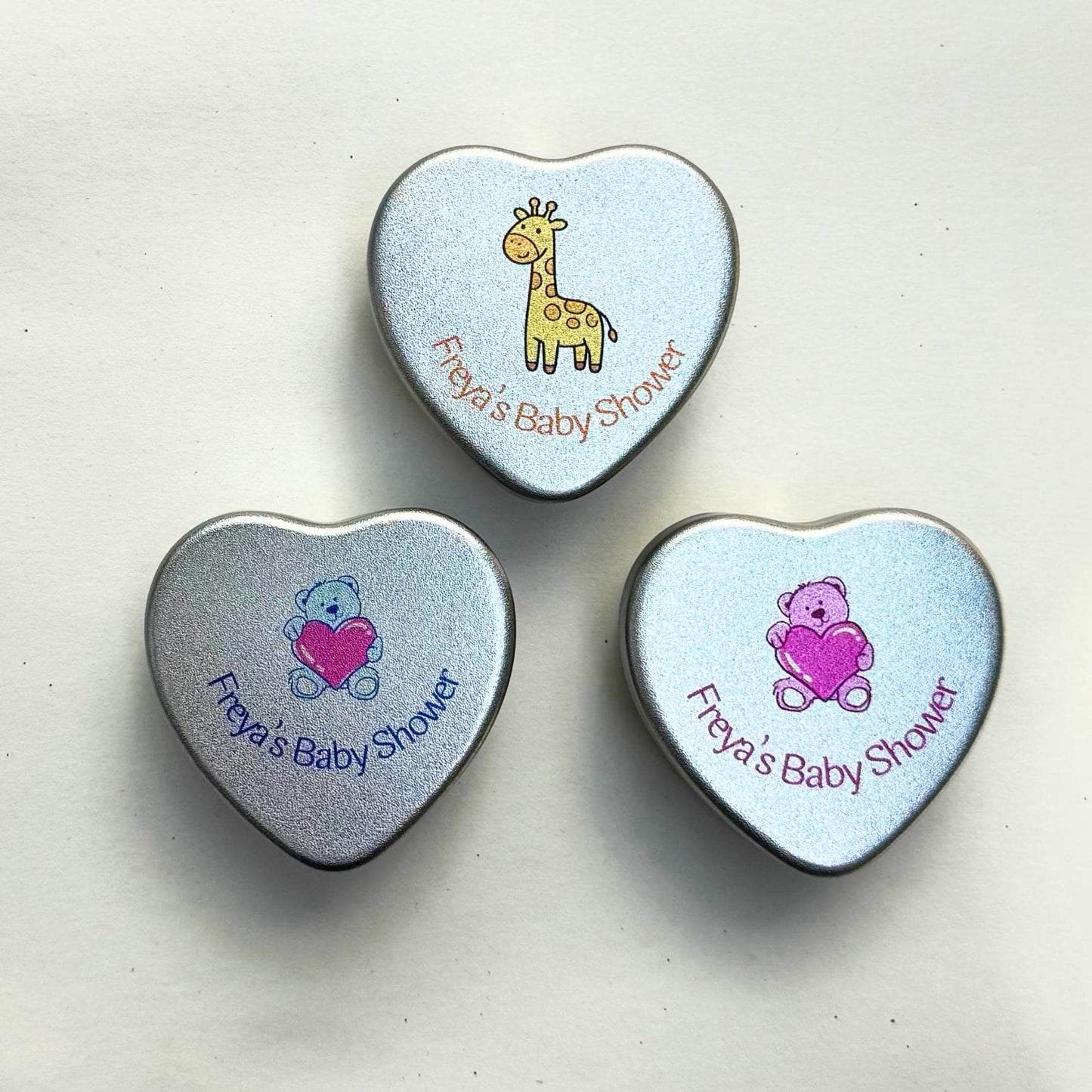 Three personalised baby shower heart tin candle favours closed, showing giraffe, blue teddy bear and pink teddy bear lid designs