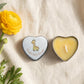 Personalised yellow baby shower candle favour — silver heart tin open showing yellow wax candle with giraffe lid design, styled with yellow ranunculus flowers