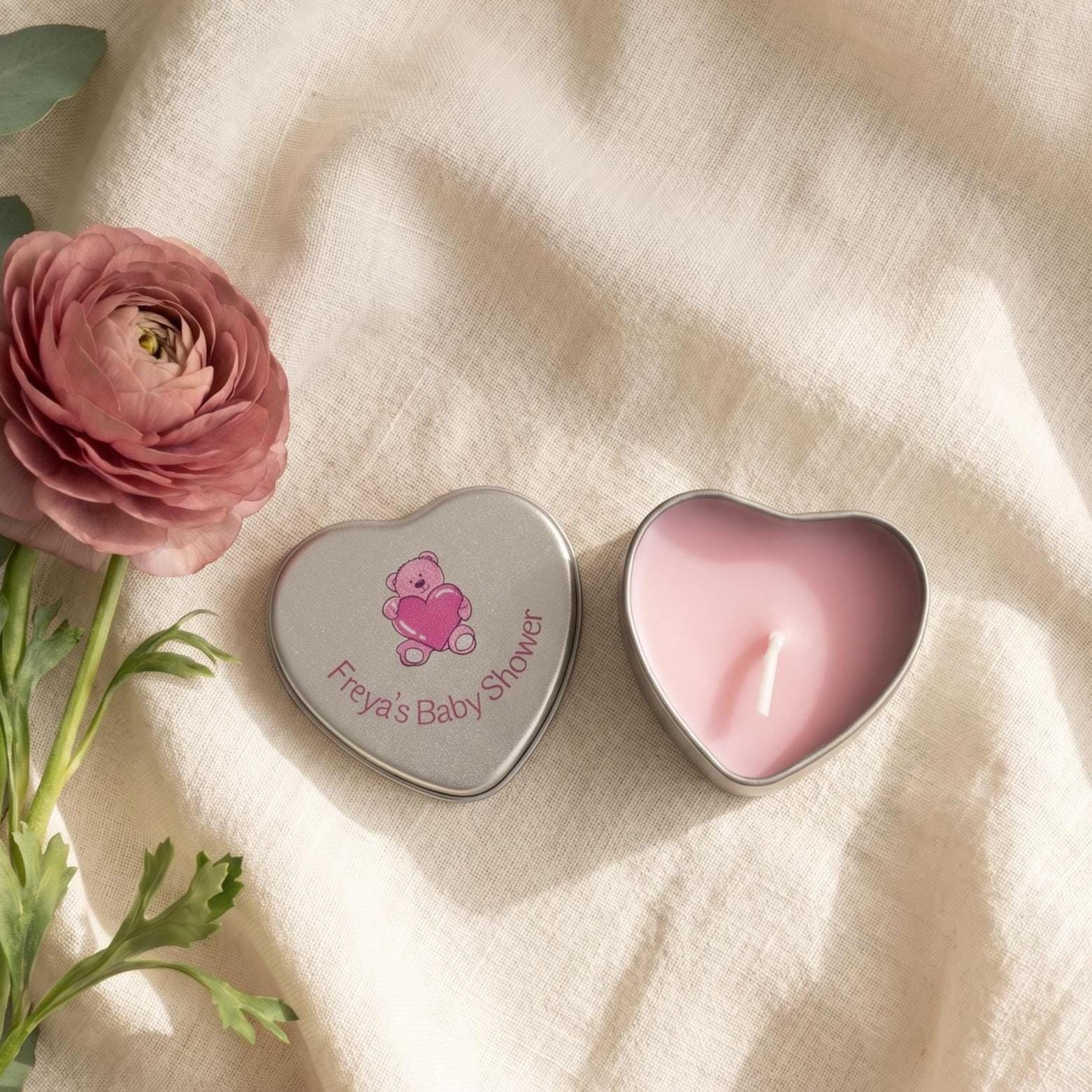 Personalised pink baby shower candle favour — silver heart tin open showing pink wax candle with teddy bear lid design, styled with pink ranunculus flowers