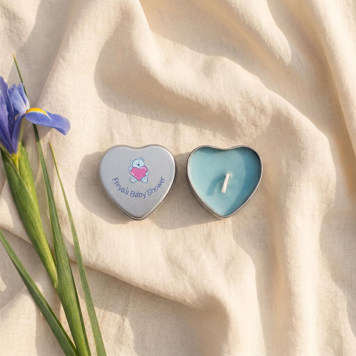 Personalised blue baby shower candle favour — silver heart tin open showing blue wax candle with teddy bear lid design, styled with iris flowers